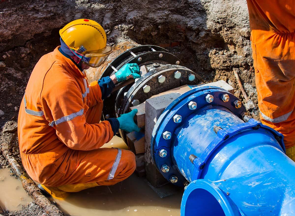 Man maintains water pipe and sits in work clothes in hole with excavated pipe.